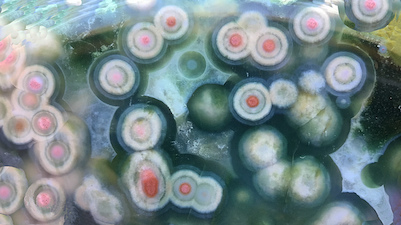 Figure 5 (Middle): Four different specimens showing spherulites 'swimming' in translucent chalcedony. The first three with clear chalcedony are likely from later collection finds, e.g. the two Marovato deposits ('veins 3/4') of 2013/14. The fourth (two closeups) with green chalcedony is from the earliest collection of translucent material, 1999/2000, and exhibits multiple ringed spherulites with vividly colorful nuclei that by eye show a random mixture of mineral particulates, e.g. iron oxide/hydroxide. Lieber's specimen in his Fig. 5 (Middle) is a quite rare green translucent with small (mm, sub-mm) round white/cream spherulites, and is a small closeup (10mm width). The colorful specimen shown here has much larger compound spherulites (5mm average). Translucent specimens with spherulites were reportedly found in early mined deposits, though due to the lack of cataloguing and documentation, we are largely unaware of just what types of translucents were mined, when and where. The colorful types span deposits 1-4 and are often smoothly 'connected' to, or interconnected with, semi-translucent or opaque varieties. I show an example of this in the Photo Gallery 16, and there are several examples in my book. Picture/specimen length: ~40mm.