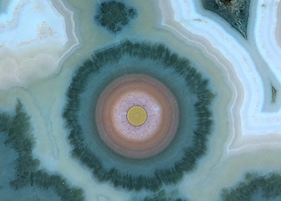 Figure 6 (Left): Macro/closeup crop of Figure 1: A compound spherulite showing inner ring structure to a core yellowish 'nucleus', surrounded by a white transition with iron oxide/hydroxide particulates, then ring structures of finer particulates, and finally bluish-green 'fibrous' growth turning into coarser dark green quartz crystals. As mentioned previously, these compound spherulites are commonly referred to as 'flower bursts' or 'flower orbs', and are quite rare, marking the difference between high grade and ultra high grade specimens. Lieber in his Fig. 6 (Left) shows a similar, but smaller and less colorful, specimen photo. Note the ringed nucleus, particulate/ring structures and outer pseudomorphic coarser crystal growth are consistent with the theory presented in my book, namely the result of silica solution spherulite nucleation, fibrous chalcedony growth mixed with 'Liesegang' type ring formation from precipitated particulates, and at later stages coarse quartz crystal growth. Picture length: ~15mm.